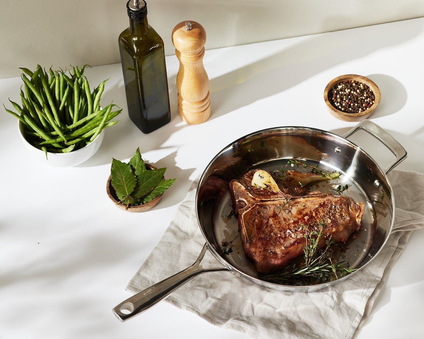 T-Bone-Steak mit Rosmarin in einer Edelstahlpfanne, daneben grüne Bohnen, Pfeffermühle und Olivenöl